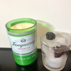 K&ouml;nnte beinhalten: Ein gr&uuml;nes Glas-Kerzengef&auml;&szlig; mit einer wei&szlig;en Kerze darin. Das Gef&auml;&szlig; tr&auml;gt den Text "Tanqueray" und "London Dry Gin". Im Hintergrund ein Glas mit Eis und Brombeeren.
