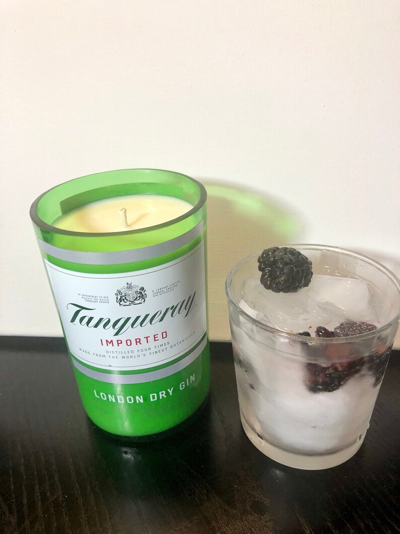 K&ouml;nnte beinhalten: Ein gr&uuml;nes Glas-Kerzengef&auml;&szlig; mit einer wei&szlig;en Kerze darin. Das Gef&auml;&szlig; tr&auml;gt den Text "Tanqueray" und "London Dry Gin". Im Hintergrund ein Glas mit Eis und Brombeeren.