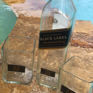 Op de afbeelding: Drie doorzichtige glazen flessen, twee zijn vierkant en &eacute;&eacute;n is rond. De ronde fles is gelabeld met "Johnnie Walker Black Label Blended Scotch Whisky". Alle drie flessen hebben een label met de tekst "Aged 12 Years".