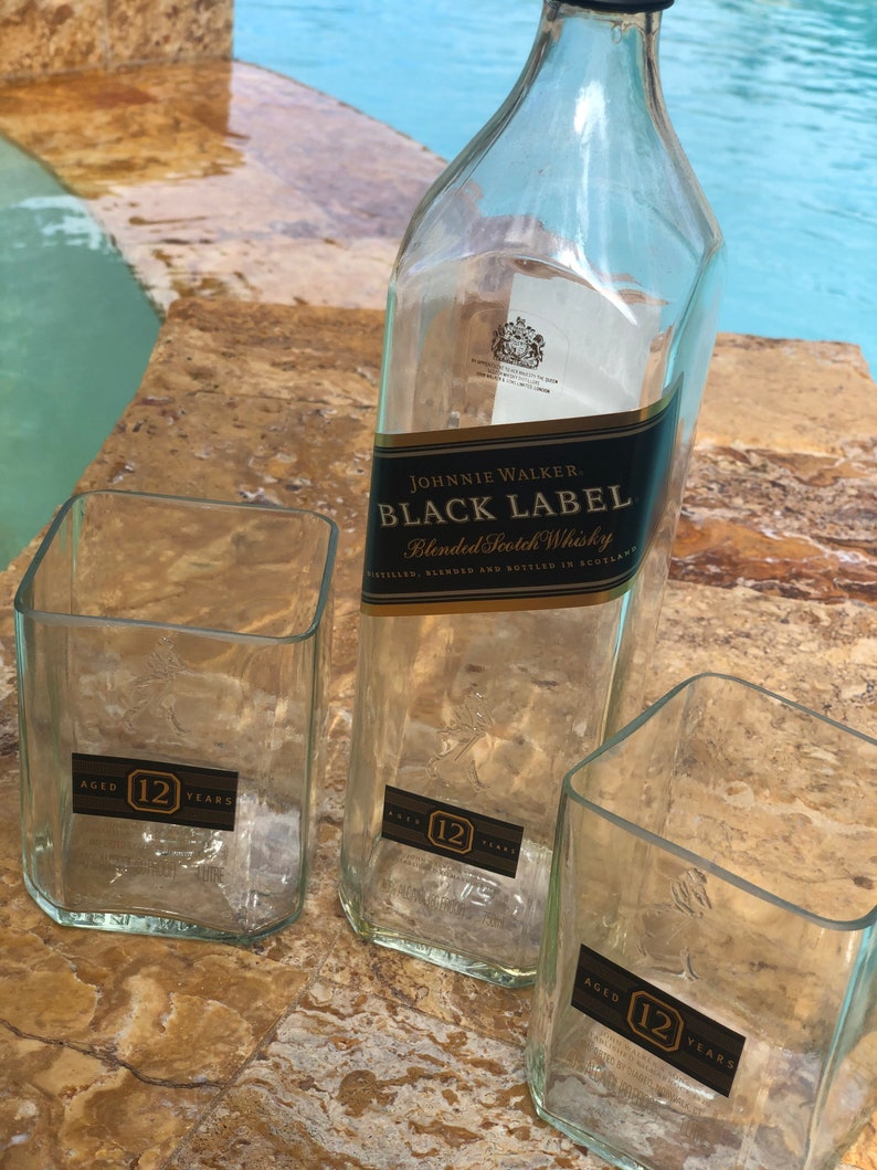Op de afbeelding: Drie doorzichtige glazen flessen, twee zijn vierkant en &eacute;&eacute;n is rond. De ronde fles is gelabeld met "Johnnie Walker Black Label Blended Scotch Whisky". Alle drie flessen hebben een label met de tekst "Aged 12 Years".