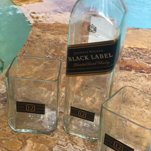Op de afbeelding: Drie doorzichtige glazen flessen met een etiket met de tekst "JOHNNIE WALKER. BLACK LABEL Blended Scotch Whisky DISTILLED, BLENDED AND BOTTLED IN SCOTLAND". Elke fles heeft een etiket met de tekst "AGED 12 YEARS".