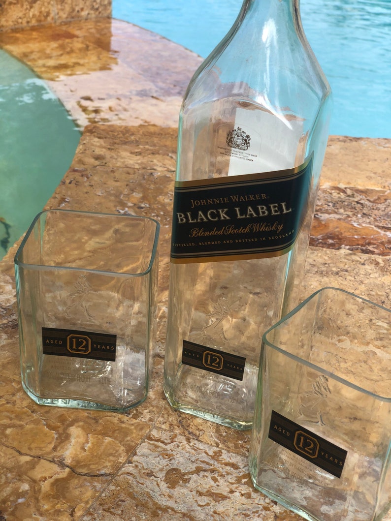Op de afbeelding: Drie doorzichtige glazen flessen met een etiket met de tekst "JOHNNIE WALKER. BLACK LABEL Blended Scotch Whisky DISTILLED, BLENDED AND BOTTLED IN SCOTLAND". Elke fles heeft een etiket met de tekst "AGED 12 YEARS".