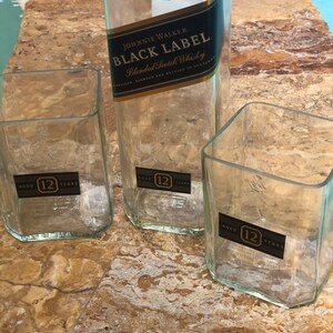 Op de afbeelding: Drie doorzichtige glazen flessen met een gouden etiket waarop "JOHNNIE WALKER. BLACK LABEL Blended Scotch Whisky" en "AGED 12 YEARS" staat.