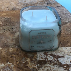 May include: A clear glass candle jar with a blue wax candle inside. The jar is labeled "SILVER PATR&Oacute;N" with the text "HECHO EN M&Eacute;XICO" and "TEQUILA, M&Eacute;XICO".