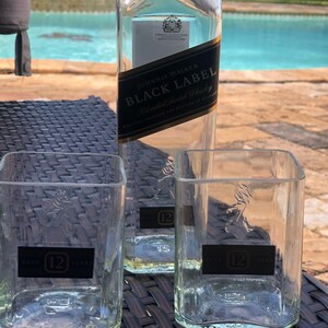 Op de afbeelding: Twee heldere glazen tumblers met het Johnnie Walker logo en "12" erop gedrukt. De tumblers staan op een zwarte rieten ondergrond. Een fles Johnnie Walker Black Label blended Scotch whisky staat op de achtergrond.