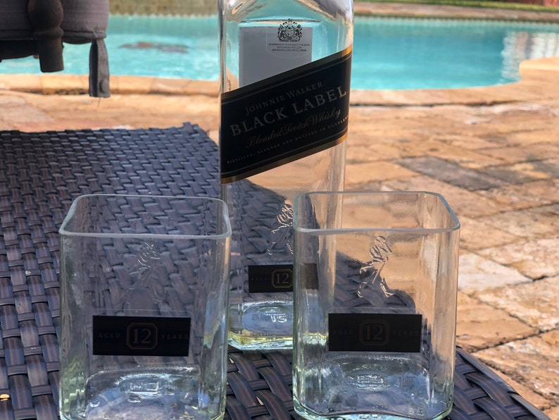 Op de afbeelding: Twee heldere glazen tumblers met het Johnnie Walker logo en "12" erop gedrukt. De tumblers staan op een zwarte rieten ondergrond. Een fles Johnnie Walker Black Label blended Scotch whisky staat op de achtergrond.