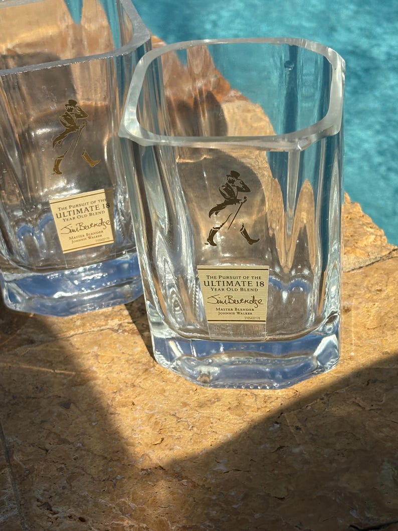 K&ouml;nnte beinhalten: Zwei klare, quadratische Schnapsgl&auml;ser mit einem Logo eines Mannes, der mit einem Stock geht. Die Gl&auml;ser tragen den Text "THE PURSUIT OF THE ULTIMATE 18 YEAR OLD BLEND" und "Sim Beverage". Sie stehen auf einer braunen Oberfl&auml;che.