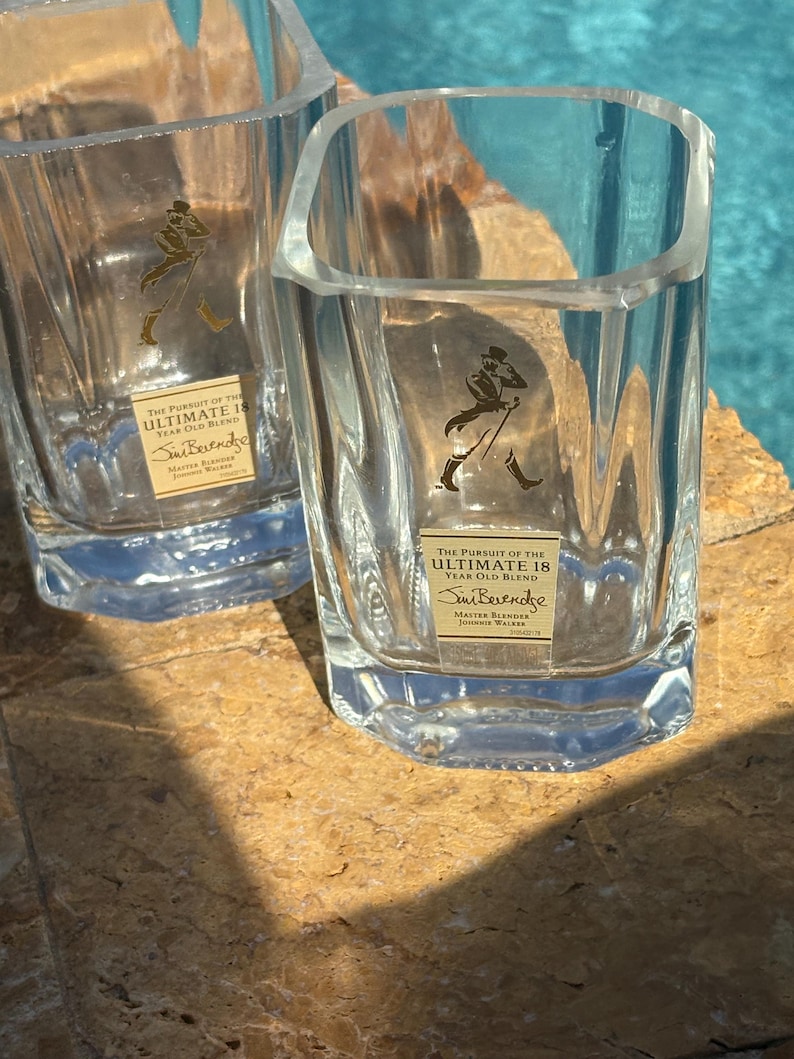K&ouml;nnte beinhalten: Zwei klare, quadratische Whiskygl&auml;ser mit Logo und Text. Die Gl&auml;ser stehen auf einer strukturierten, hellbraunen Oberfl&auml;che mit blauem Hintergrund. Der Text auf den Gl&auml;sern lautet "THE PURSUIT OF THE ULTIMATE 18 YEAR OLD BLEND".