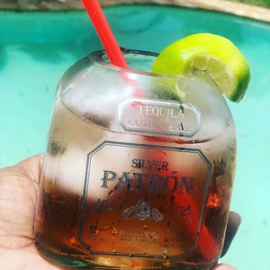 May include: A glass filled with a clear liquid and a lime wedge, with a red straw sticking out. The glass is made from a recycled Patron tequila bottle. The bottle label reads "Tequila 100% de Agave Silver Patron Hecho en Mexico Made in Mexico 40% Alc./Vol. 80 Proof."