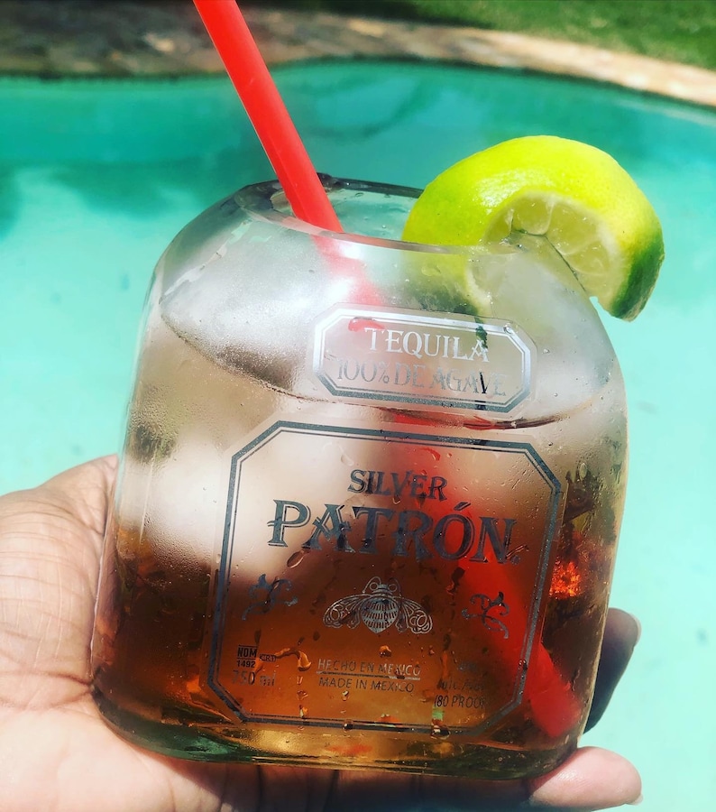 May include: A glass filled with a clear liquid and a lime wedge, with a red straw sticking out. The glass is made from a recycled Patron tequila bottle. The bottle label reads "Tequila 100% de Agave Silver Patron Hecho en Mexico Made in Mexico 40% Alc./Vol. 80 Proof."