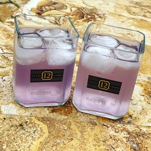 Op de afbeelding: Twee heldere glazen bekers gevuld met een lichtpaarse vloeistof en ijsblokjes. De bekers hebben etiketten met de tekst "Aged 12 Years John Walker & Sons Established Kilmarnock 1820 40% Alc/Vol (80 Proof) 750ml".