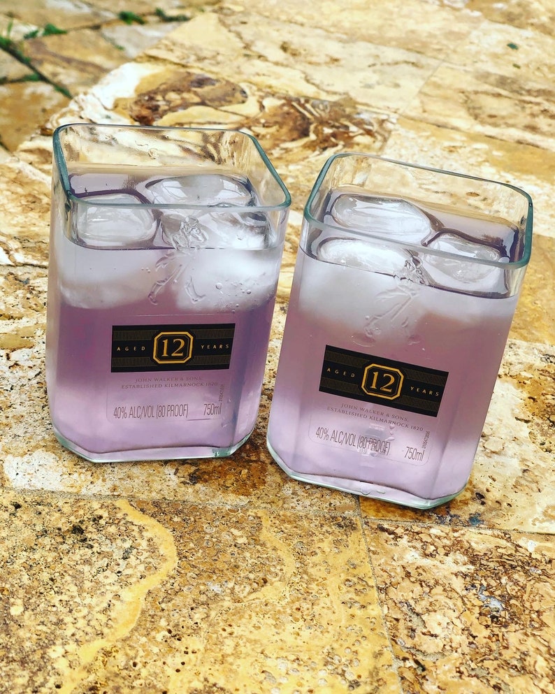 Op de afbeelding: Twee heldere glazen bekers gevuld met een lichtpaarse vloeistof en ijsblokjes. De bekers hebben etiketten met de tekst "Aged 12 Years John Walker & Sons Established Kilmarnock 1820 40% Alc/Vol (80 Proof) 750ml".