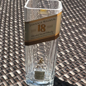 May include: A clear glass bottle with a gold label that reads "JOHNNIE WALKER AGED 18 YEARS Blended Scotch Whisky". The bottle is sitting on a brown wicker surface.