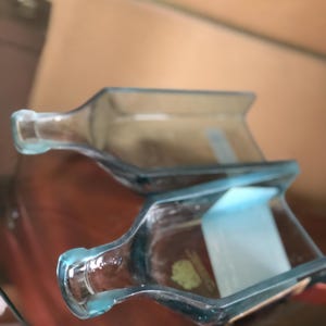 以下が含まれることがあります： 2つの透明なガラス製のボトル型トレイ。トレイは、わずかに青みがかった透明なガラスでできています。トレイは茶色の表面に置かれています。トレイは、ボトルの首が付いた長方形の形をしています。