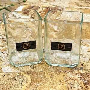 Op de afbeelding: Twee heldere glazen bekers gemaakt van hergebruikte 12 jaar oude John Walker & Sons whiskyflessen. Elke beker heeft een etiket met de tekst "AGED 12 YEARS" en "40% ALC/VOL 80 PROOF 750ml".