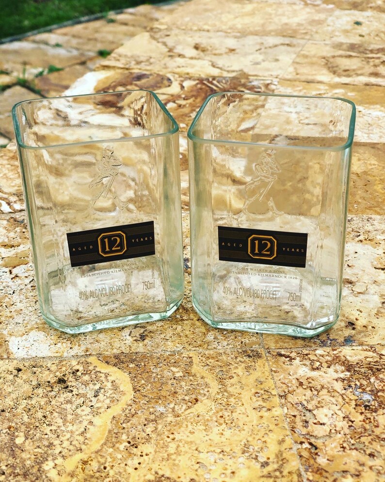 Op de afbeelding: Twee heldere glazen bekers gemaakt van hergebruikte 12 jaar oude John Walker & Sons whiskyflessen. Elke beker heeft een etiket met de tekst "AGED 12 YEARS" en "40% ALC/VOL 80 PROOF 750ml".