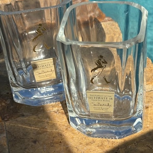 K&ouml;nnte beinhalten: Zwei klare, quadratische Whiskygl&auml;ser mit facettiertem Design. Jedes Glas hat ein goldenes Etikett mit Text und einer Figur. Der Text auf dem Etikett lautet "THE PURSUIT OF THE ULTIMATE 18 YEAR OLD BLEND" und "Sim Berendse".