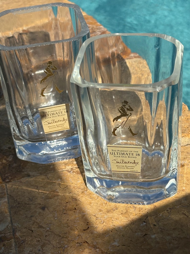 K&ouml;nnte beinhalten: Zwei klare, quadratische Whiskygl&auml;ser mit facettiertem Design. Jedes Glas hat ein goldenes Etikett mit Text und einer Figur. Der Text auf dem Etikett lautet "THE PURSUIT OF THE ULTIMATE 18 YEAR OLD BLEND" und "Sim Berendse".