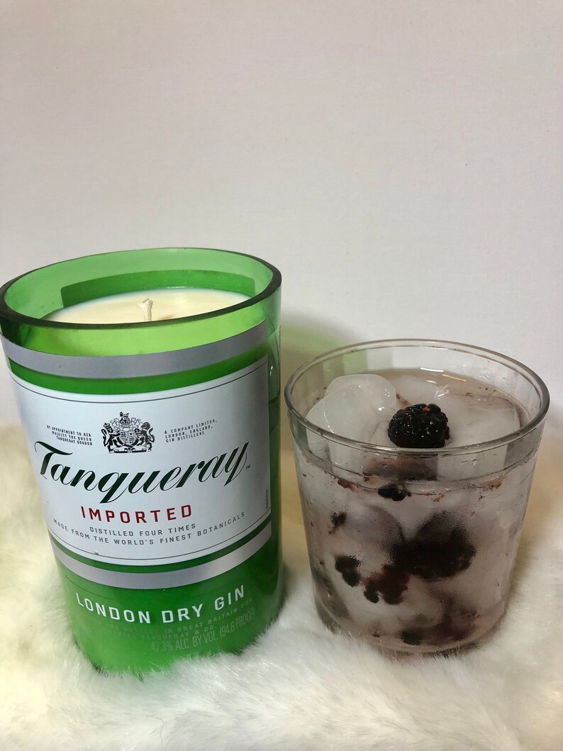 K&ouml;nnte beinhalten: Ein gr&uuml;nes Glas-Kerzengef&auml;&szlig; mit einer wei&szlig;en Kerze darin. Das Gef&auml;&szlig; ist mit "Tanqueray Imported London Dry Gin" beschriftet und tr&auml;gt ein k&ouml;nigliches Wappen auf dem Etikett. Im Hintergrund steht ein Glas mit Eiswasser und Brombeeren.