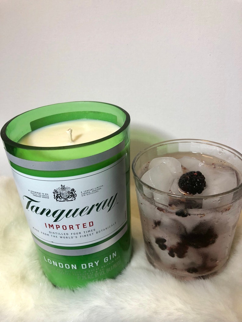 K&ouml;nnte beinhalten: Ein gr&uuml;nes Glas-Kerzengef&auml;&szlig; mit einer wei&szlig;en Kerze darin. Das Gef&auml;&szlig; ist mit "Tanqueray Imported London Dry Gin" beschriftet. Im Hintergrund ein Glas mit Eis und Brombeeren.