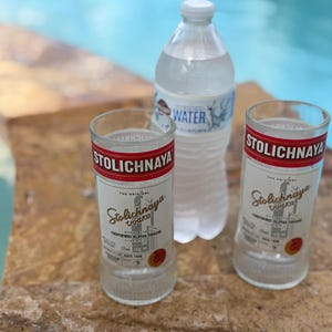 以下が含まれることがあります： 赤と白のラベルが付いた2つの空のストリチナヤウォッカグラスと、青と白のラベルが付いた透明なプラスチックウォーターボトル。アイテムはプールの近くの石の表面にあります。