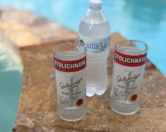 May include: Two empty Stolichnaya vodka glasses with red and white labels, and a clear plastic water bottle with a blue and white label. The items are on a stone surface near a pool.