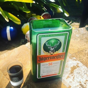 May include: A green candle in a repurposed Jägermeister bottle, with the brand's label visible. A small shot glass with a dark liquid sits beside it. The candle has a light green wax and a wick.