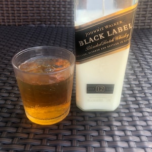 May include: A clear glass candle in the shape of a Johnnie Walker Black Label Scotch Whisky bottle. The candle is white and has a black label with gold lettering that reads "Johnnie Walker Black Label Blended Scotch Whisky Distilled, Blended and Bottled in Scotland Aged 12 Years".