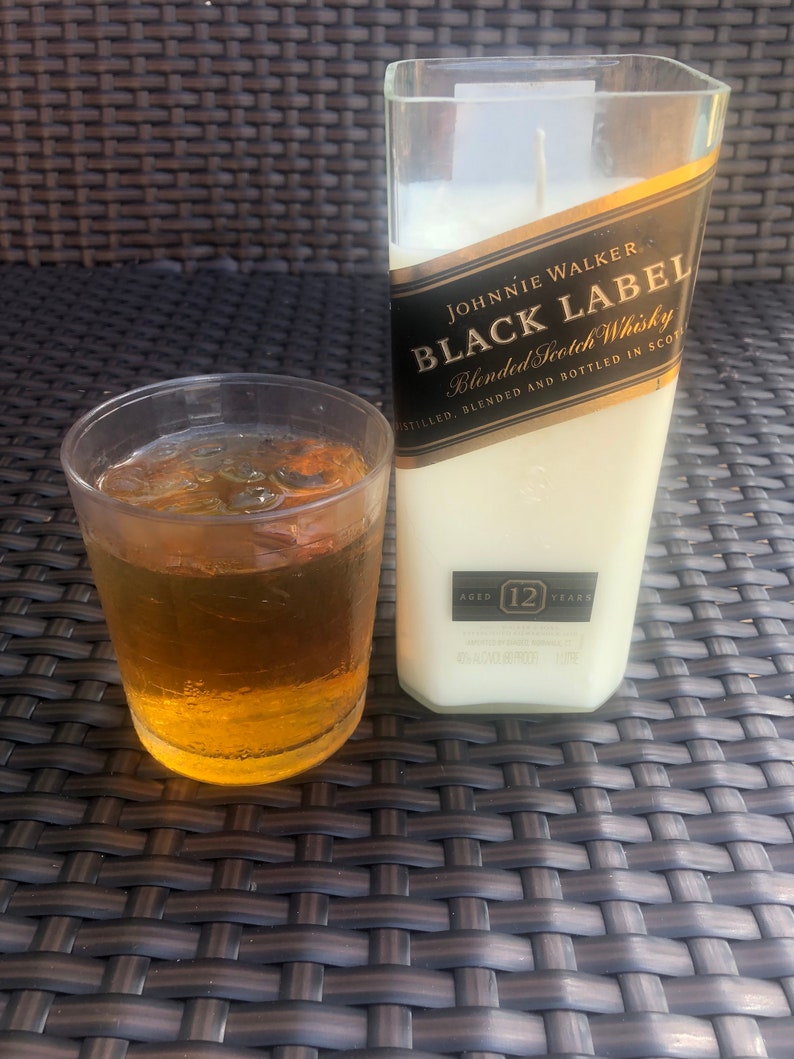 May include: A clear glass candle in the shape of a Johnnie Walker Black Label Scotch Whisky bottle. The candle is white and has a black label with gold lettering that reads "Johnnie Walker Black Label Blended Scotch Whisky Distilled, Blended and Bottled in Scotland Aged 12 Years".