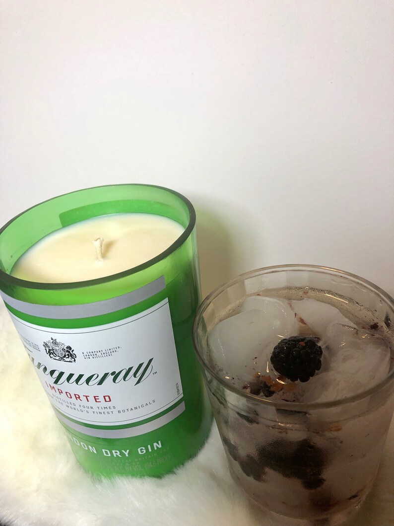 K&ouml;nnte beinhalten: Ein gr&uuml;nes Glas-Kerzengef&auml;&szlig; mit einer wei&szlig;en Kerze darin. Das Gef&auml;&szlig; tr&auml;gt ein Etikett mit der Aufschrift "Tanqueray Importiert" und "London Dry Gin". Neben der Kerze steht ein Glas mit Eis und einer Brombeere.