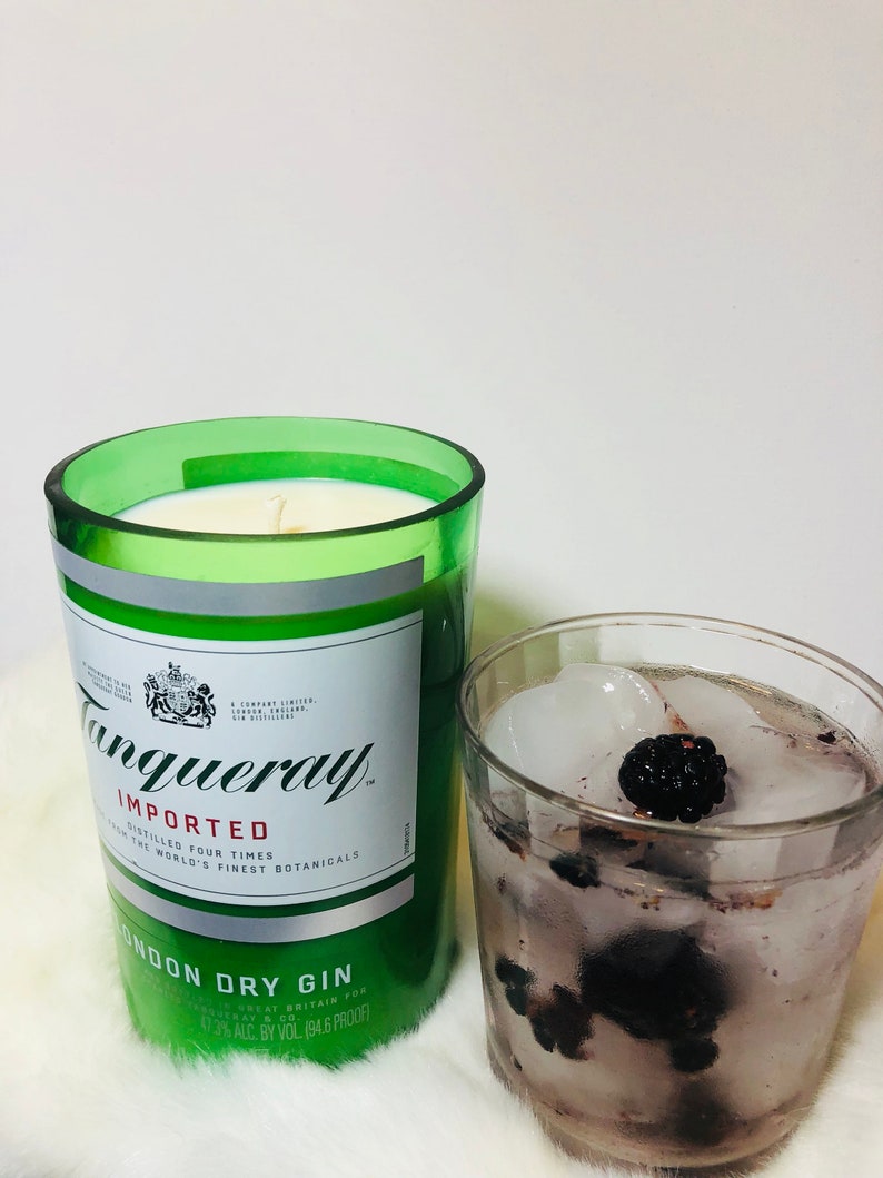 K&ouml;nnte beinhalten: Ein gr&uuml;nes Glas-Kerzengef&auml;&szlig; mit einem wei&szlig;en Etikett, auf dem "Tanqueray Imported" und "London Dry Gin" steht. Das Kerzengef&auml;&szlig; steht auf einer wei&szlig;en Oberfl&auml;che neben einem Glas mit Eis und Brombeeren.