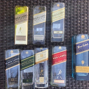 May include: A collection of eight empty glass bottles of Johnnie Walker Scotch whisky. The bottles are arranged in two rows of four. The bottles are all different sizes and shapes, and they have different labels. The labels include the Johnnie Walker logo, the brand name, the age of the whisky, and the type of whisky. The bottles are all clear glass and have a slightly frosted appearance.