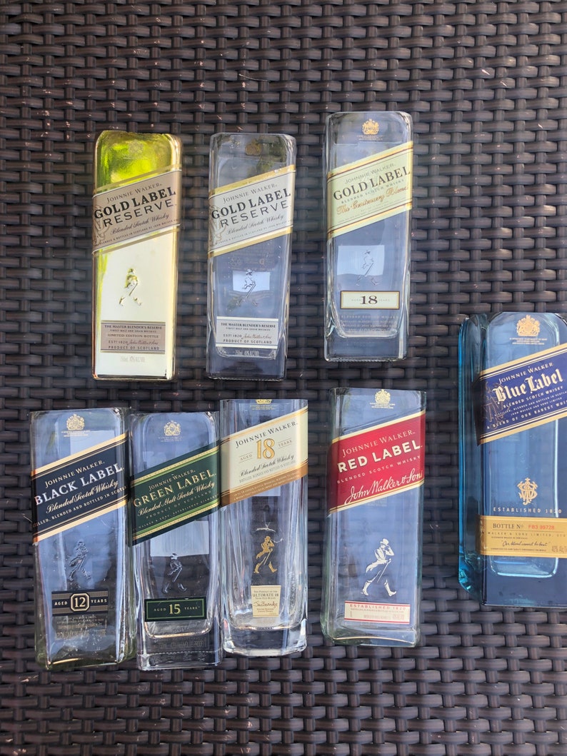 May include: A collection of eight empty glass bottles of Johnnie Walker Scotch whisky. The bottles are arranged in two rows of four. The bottles are all different sizes and shapes, and they have different labels. The labels include the Johnnie Walker logo, the brand name, the age of the whisky, and the type of whisky. The bottles are all clear glass and have a slightly frosted appearance.
