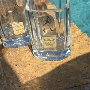 K&ouml;nnte beinhalten: Zwei klare, quadratische Whiskygl&auml;ser mit dem Text "THE PURSUIT OF THE ULTIMATE 18 YEAR OLD BLEND" und "Sim Berendse" auf einem goldenen Etikett. Die Gl&auml;ser stehen auf einer hellbraunen Oberfl&auml;che.