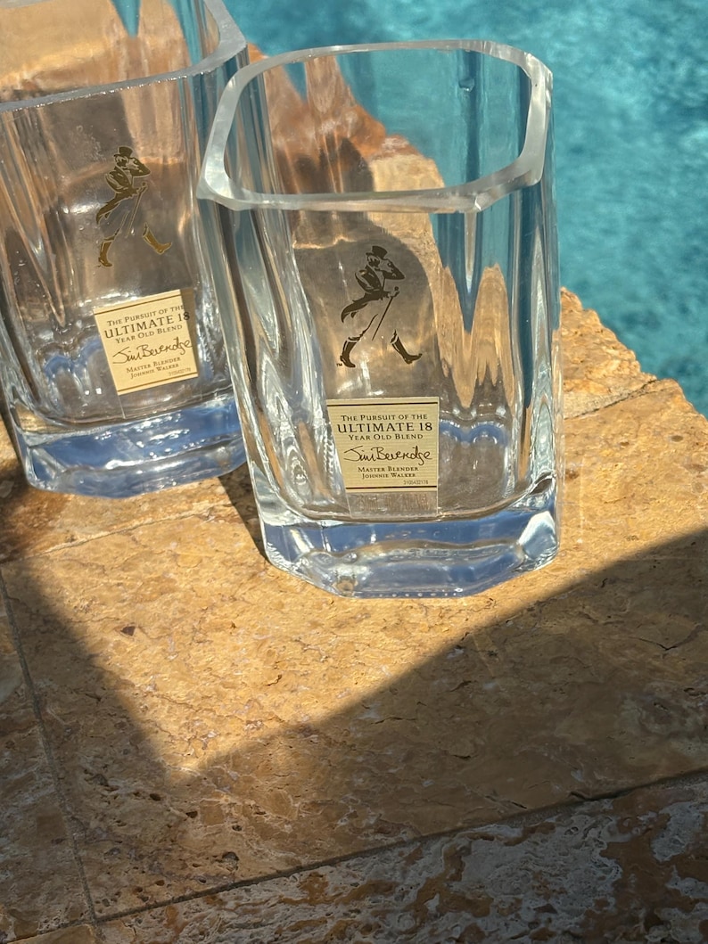 K&ouml;nnte beinhalten: Zwei klare, quadratische Whiskygl&auml;ser mit dem Text "THE PURSUIT OF THE ULTIMATE 18 YEAR OLD BLEND" und "Sim Berendse" auf einem goldenen Etikett. Die Gl&auml;ser stehen auf einer hellbraunen Oberfl&auml;che.