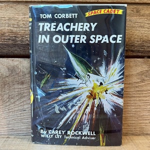 May include: A vintage paperback book cover with a colorful illustration of a spaceship exploding in outer space. The title is "Treachery in Outer Space" by Carey Rockwell. The book is part of the "Space Cadet" series by Tom Corbett.