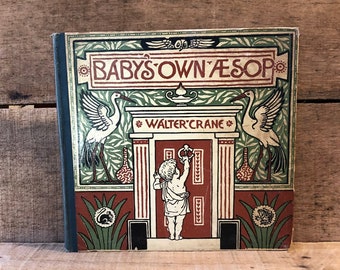 May include: A vintage book titled "Baby's Own Aesop" by Walter Crane. The cover features a child in a white tunic standing in a doorway with two white cranes on either side. The book has a green, red, and gold design.