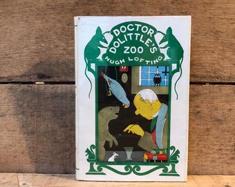 Puede incluir: Un libro antiguo de tapa dura titulado "Doctor Dolittle's Zoo" de Hugh Lofting. La portada presenta un fondo blanco con un borde verde e ilustraciones de un hombre, un loro, un ratón y un tejón.