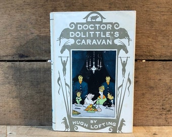 May include: A white book with a green and gray illustration of a pig and a group of people sitting at a table. The title of the book is "Doctor Dolittle's Caravan" by Hugh Lofting.