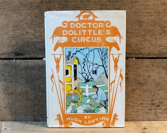 May include: A vintage book cover with a white background and orange accents. The title is "Doctor Dolittle's Circus" by Hugh Lofting. The cover features an illustration of a circus wagon with animals and a man in a blue shirt.