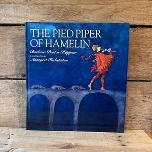 May include: A blue hardcover book titled "The Pied Piper of Hamelin" by Barbara Bartos-Höppner, illustrated by Annegert Fuchshuber. The cover features a colorful illustration of a man playing a pipe, with rats following him across a bridge.