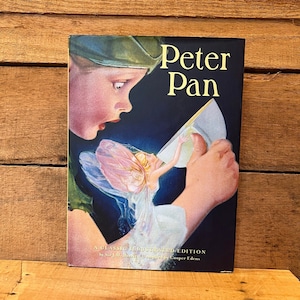 May include: A hardcover book titled "Peter Pan" with an illustration of a boy in a green hat looking at a fairy. The book cover has the text "Peter Pan" in gold lettering. The book is a classic illustrated edition.