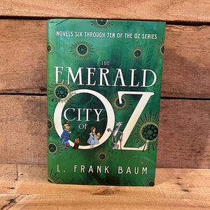 May include: A green hardcover book with a title that reads "The Emerald City of Oz" and the author's name "L. Frank Baum". The cover features illustrations of characters from the book, including the Scarecrow, Dorothy, and the Tin Man.