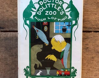 May include: A hardback book titled Doctor Dolittle's Zoo by Hugh Lofting with a white dust jacket featuring a green border, two rat silhouettes, and a central illustration of a man interacting with a parrot, an otter, a mouse, and an owl.
