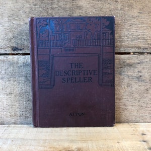 May include: A vintage hardcover book titled "The Descriptive Speller" with a dark burgundy cover. The cover features a black illustration of a house and the author's name, Aiton. The book is displayed on a wooden surface.