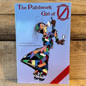 May include: A book titled "The Patchwork Girl of Oz" by L. Frank Baum, illustrated by John R. Neill. The cover features a colorful illustration of a patchwork doll-like character holding a magnifying glass and a piece of paper, set against a light blue background.