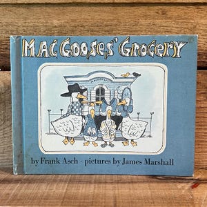 May include: A children's book titled "Mac Gooses' Grocery" with a light blue cover. The cover illustration features cartoon geese in front of a building. The author is Frank Asch, and the pictures are by James Marshall.