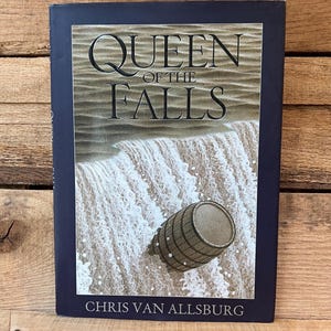 May include: A hardcover book titled "QUEEN OF THE FALLS" by Chris Van Allsburg. The cover features a waterfall scene with a wooden barrel. The book has a dark blue border and is set against a wooden background.