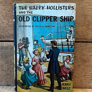 May include: A hardcover book titled The Happy Hollisters and the Old Clipper Ship by Jerry West, illustrated by Helen S. Hamilton. The cover features a colorful illustration of people on a ship deck, including a person filming with a movie camera.