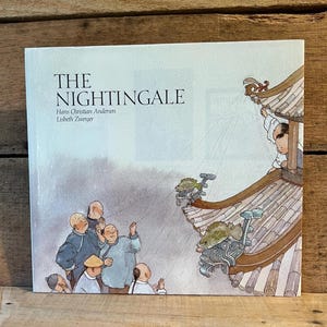 May include: A hardcover book titled "The Nightingale" by Hans Christian Andersen and Lisbeth Zwerger. The cover features an illustration of people looking up at a building with ornate roof details and figures. The color palette includes blues, browns, and whites.